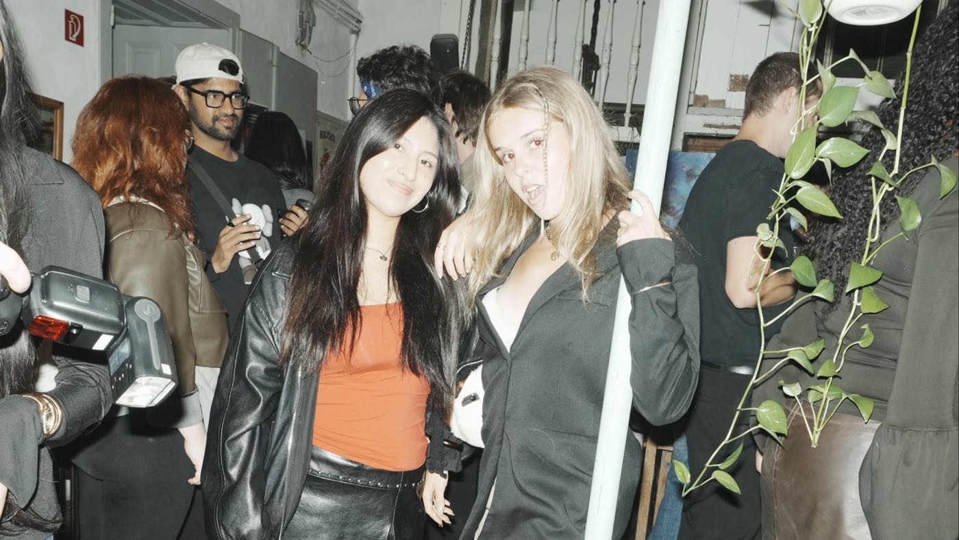 Two guests posing together beside a metal pole in a busy room, other guests mingling behind them and a photographer with a flash at the left edge.