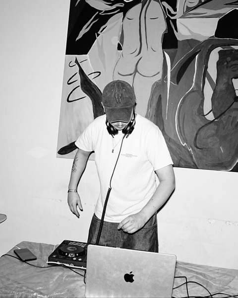A DJ in a cap and headphones playing from decks and a laptop in front of a large painting.