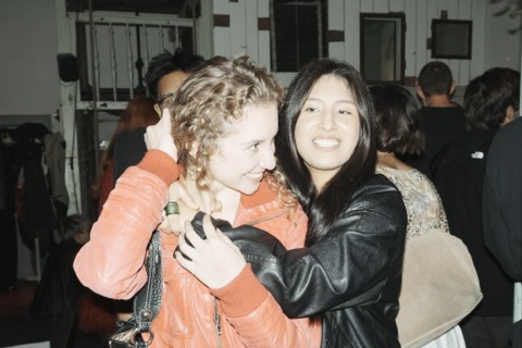 Two guests hugging and laughing, one in an orange leather jacket and one in black, with a busy room behind them.