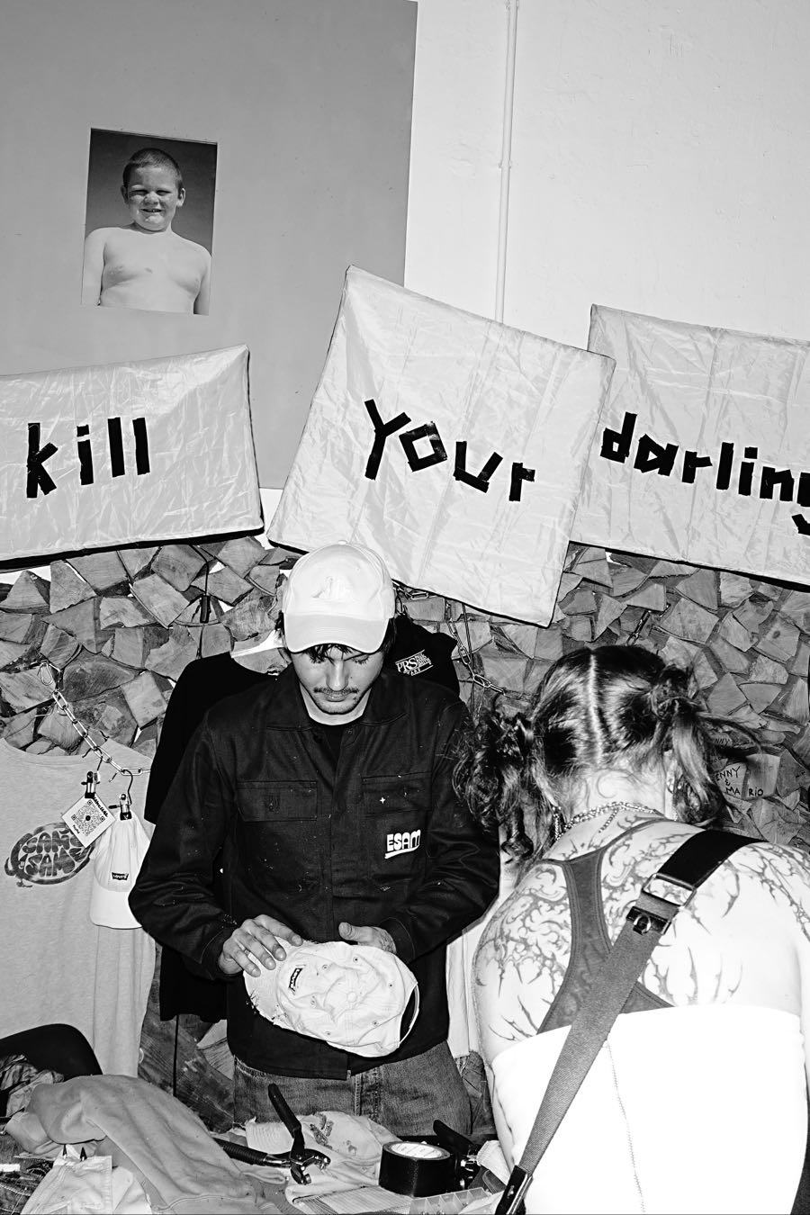 A stall seller in a dark work jacket folding a cap beneath hand-lettered banners that read kill your darling, with a customer in the foreground.