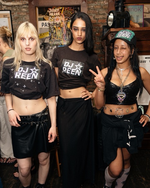 Three guests in black tops and skirts posing side by side, one making a peace sign, in front of a wall of posters.