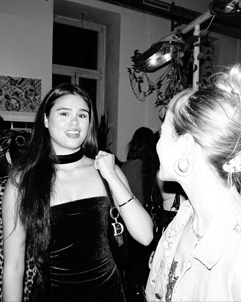 A guest in a black velvet dress and choker smiling at another guest seen in profile, a busy room behind them.